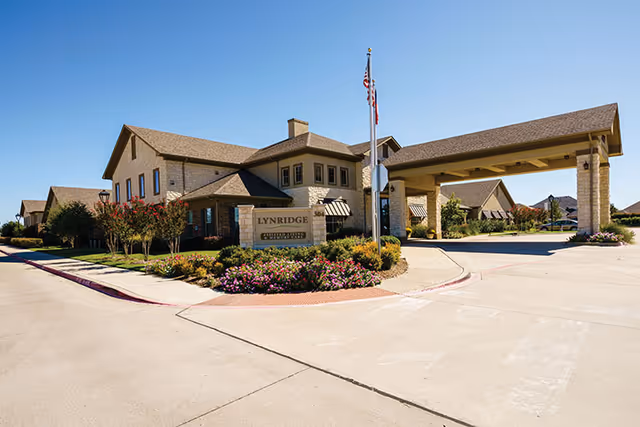 Exterior view of Lynridge of Murphy Assisted Living & Memory Care facility showing a large building with a covered entrance, landscaped flower beds, and an American flag on a flagpole under a clear blue sky.