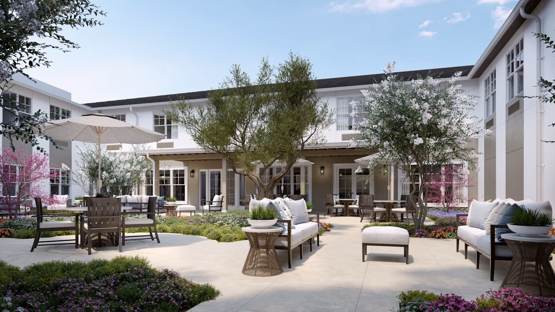 Outdoor courtyard area at Sunrise of Oceanside featuring comfortable seating with cushions, tables with umbrellas, flowering trees, and well-maintained landscaping surrounded by a two-story building under a clear sky.