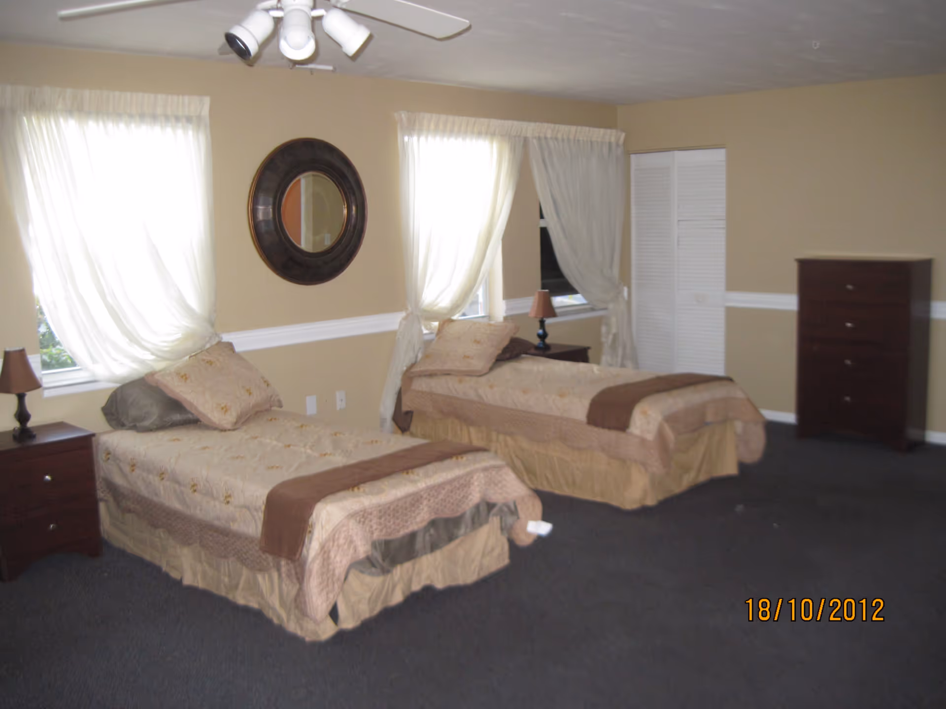 A bedroom with two single beds, each with beige and brown bedding. There are two windows with white curtains behind the beds, a round mirror on the wall between the windows, two bedside tables with lamps, a white closet door, and a tall dark wooden dresser against the beige walls.