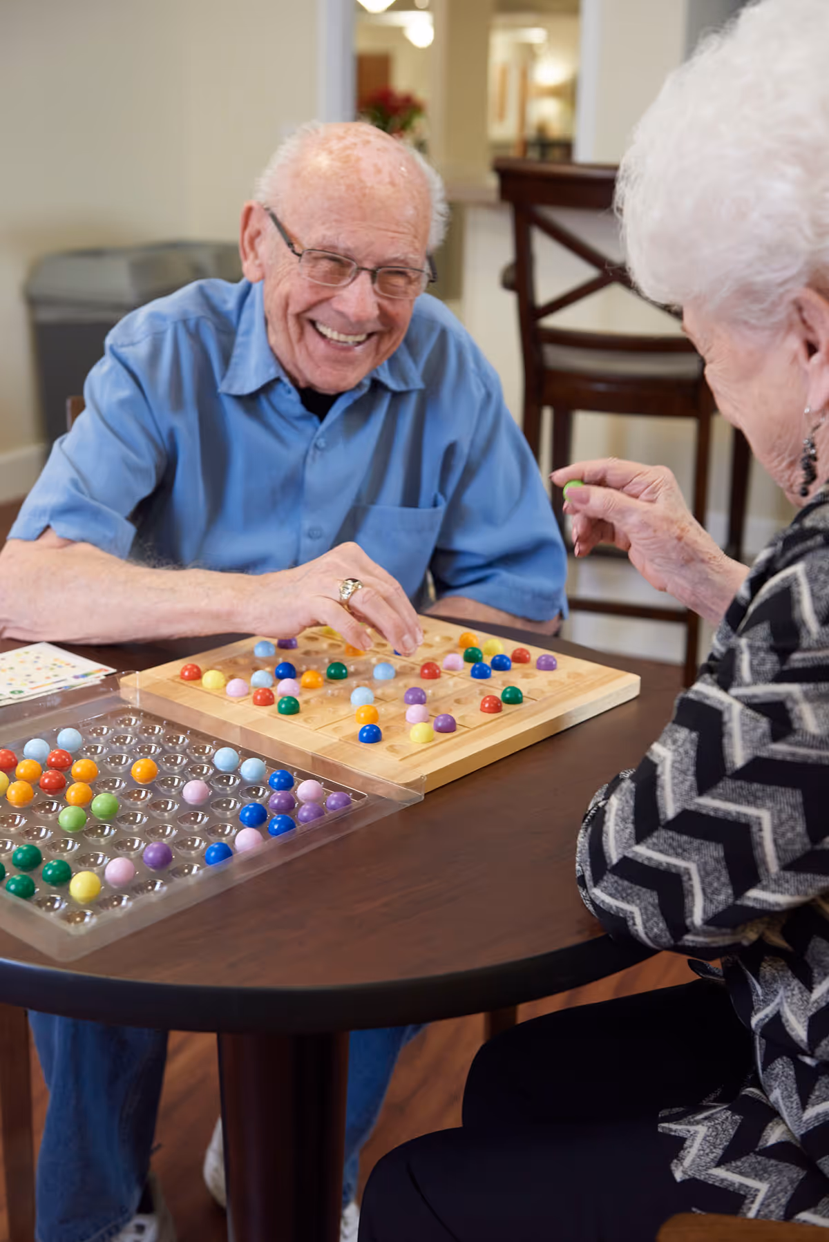 An elderly man and woman sitting at a round table playing a colorful marble board game together indoors. The man is smiling and wearing glasses and a blue shirt, while the woman is holding a green marble and wearing a patterned sweater.