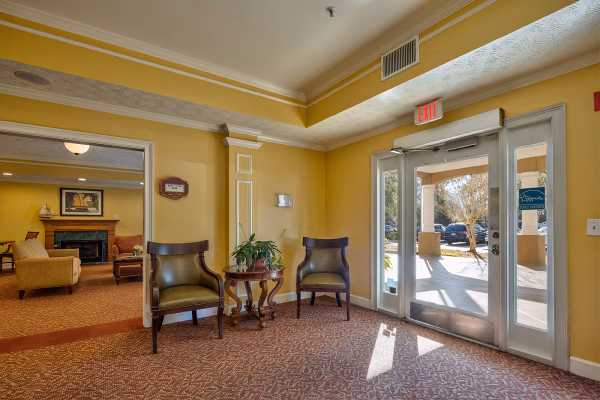 Interior view of a senior living facility entrance area with yellow walls, two green cushioned chairs flanking a small wooden table with a plant, and a glass door leading outside. Through an open doorway, a cozy living room with a fireplace, beige and orange seating, and a framed picture on the wall is visible.