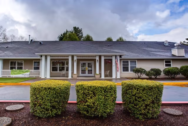 Front exterior view of a single-story building with a covered entrance, three neatly trimmed bushes in the foreground, an American flag hanging near the entrance, and a green sign on the left side indicating the facility name Grace Place.