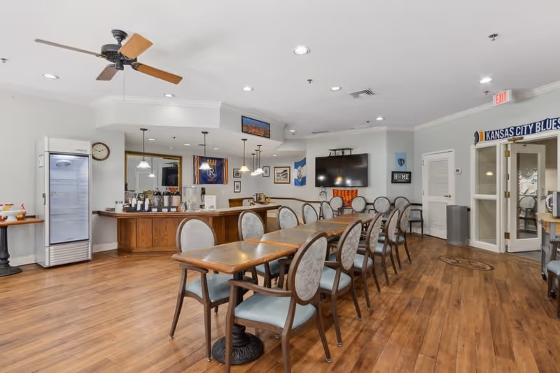 A spacious dining room with a long wooden table surrounded by cushioned chairs. The room features wood flooring, a ceiling fan, recessed lighting, a large flat-screen TV mounted on the wall, and a beverage station with coffee and water dispensers. There are various decorations on the walls, including flags and framed pictures, and double glass doors leading to another area.