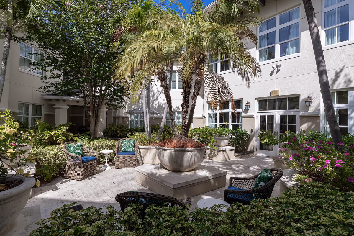 Landscaped courtyard with palm trees, wicker chairs, large planters and a white multi-story building with windows.