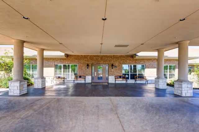 Covered entrance area of a building with four stone pillars supporting the roof. There are four benches placed against a brick wall with two large windows and a double glass door in the center. The ground is paved with concrete.