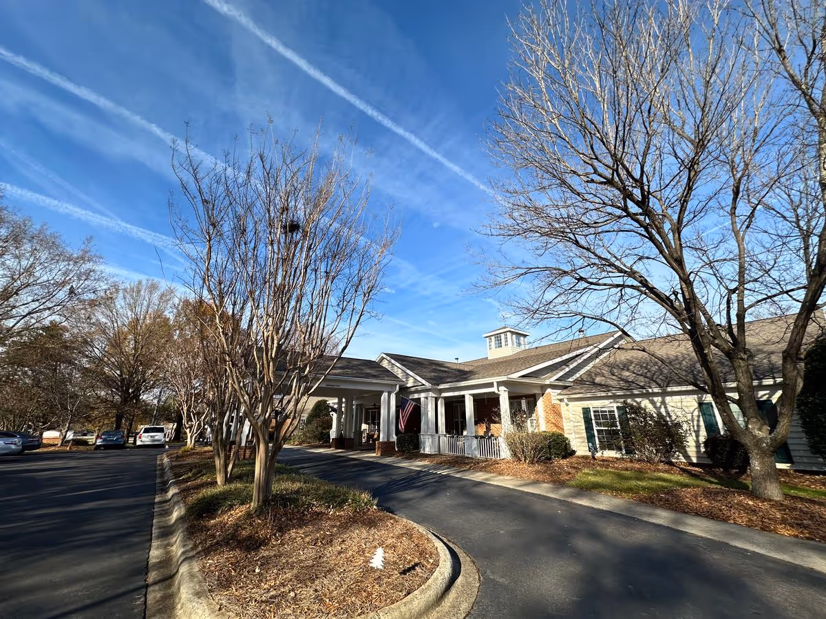 Front entrance of Chandler Place Independent Living showing a one-story building with a covered portico, bare trees, parked cars, and a blue sky with contrails.
