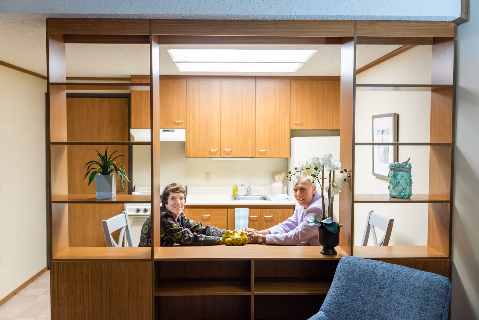 Two elderly people sitting at a kitchen table in a retirement village kitchen, visible through a wooden shelving partition. The kitchen has wooden cabinets, a white refrigerator, and a stove. There is a potted orchid on the counter and a blue chair in the foreground.