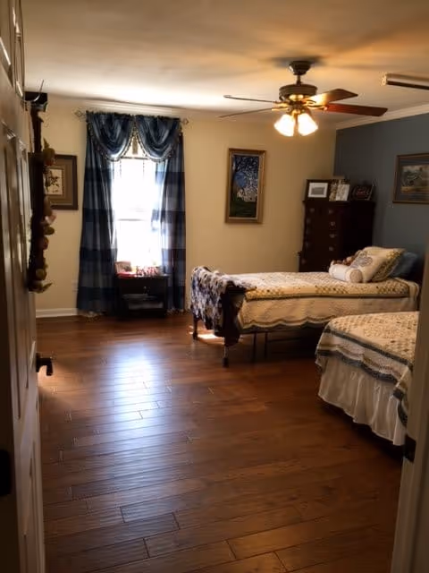 A cozy bedroom with two twin beds covered in patterned quilts. The room has wooden flooring, a ceiling fan with lights, a window with blue plaid curtains, a small nightstand with items on it, and framed artwork on the walls. There is also a tall dresser with picture frames on top.