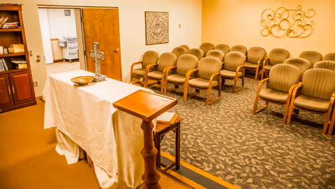 Small chapel-style meeting room with an altar draped in a white cloth, a cross and podium up front, and rows of upholstered chairs.