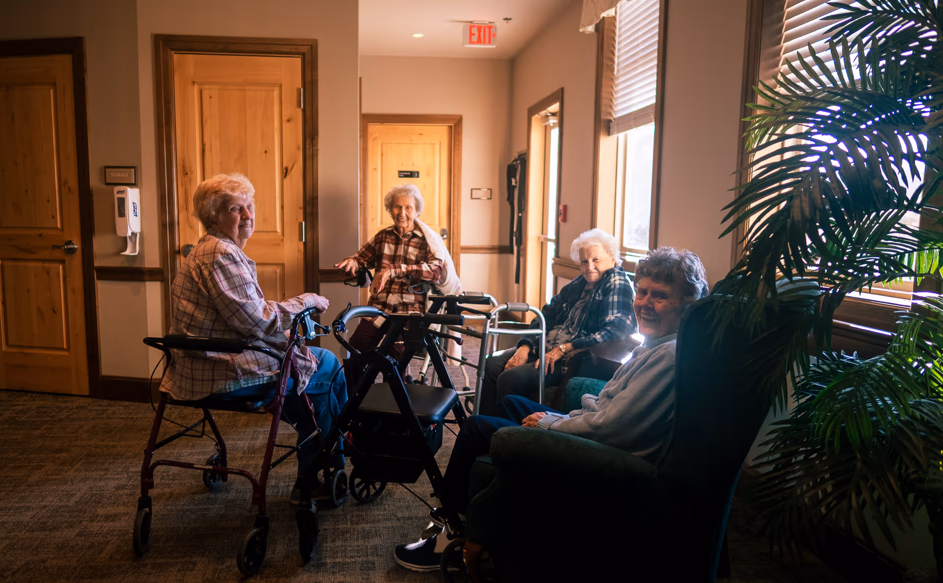 Four elderly women sitting and chatting in a well-lit hallway of a senior living facility. Two women are seated in chairs near a window with a large green plant beside them, while the other two women are using walkers. Wooden doors and an exit sign are visible in the background.
