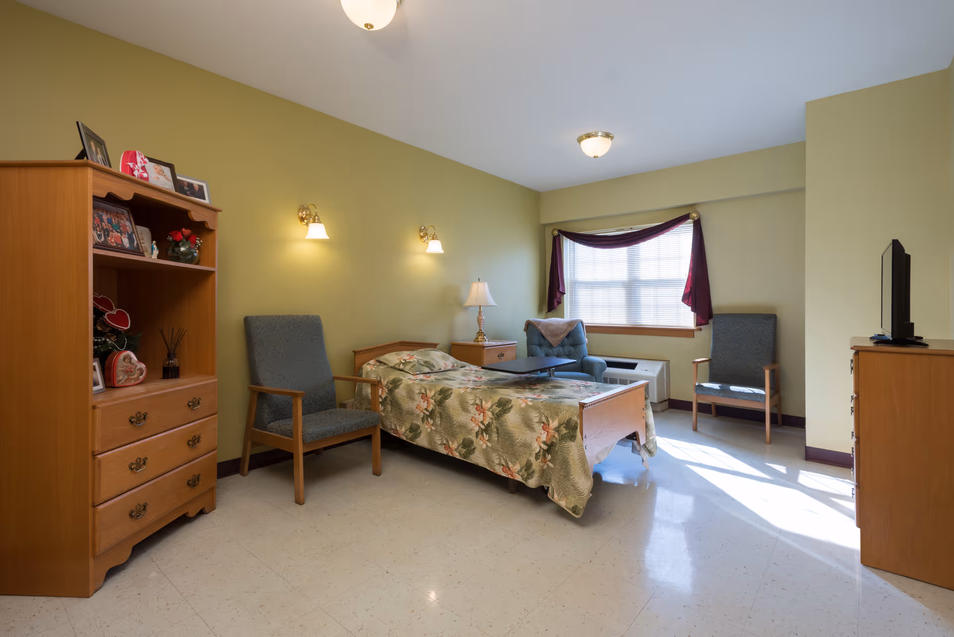 A well-lit bedroom in a senior living facility with a single bed covered in a floral bedspread, two armchairs, a wooden dresser with a TV on top, a wooden shelving unit with framed photos and decorations, and a window with burgundy curtains letting in natural light.
