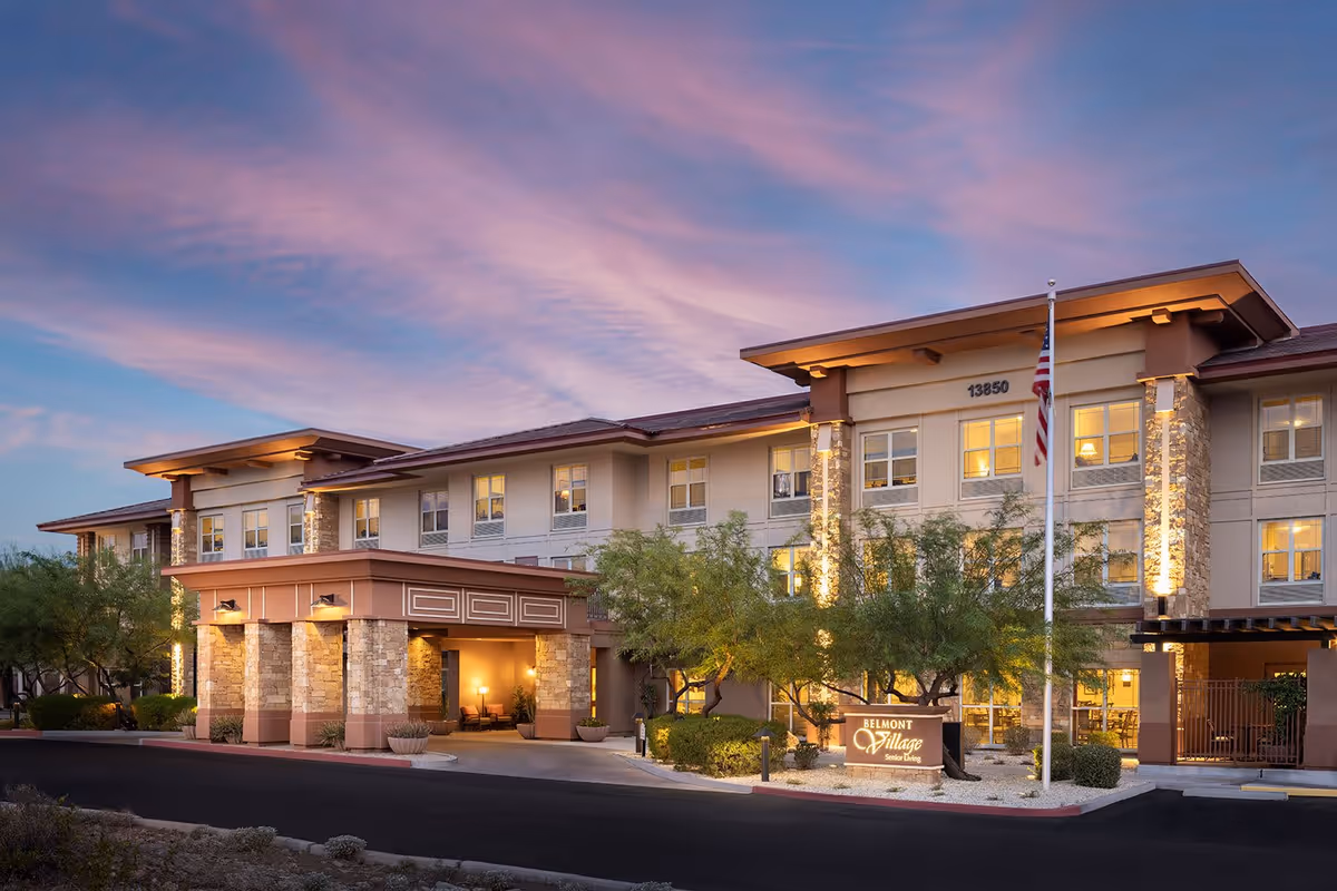 Exterior view of Belmont Village Senior Living Scottsdale building at dusk with warm lights glowing from the windows, a covered entrance with stone pillars, trees and landscaping in front, and an American flag on a flagpole.