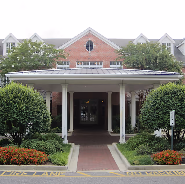 Front entrance of a senior living facility with a covered driveway supported by white columns, surrounded by neatly trimmed bushes and flower beds. The building has a brick facade with multiple windows and a gray roof.