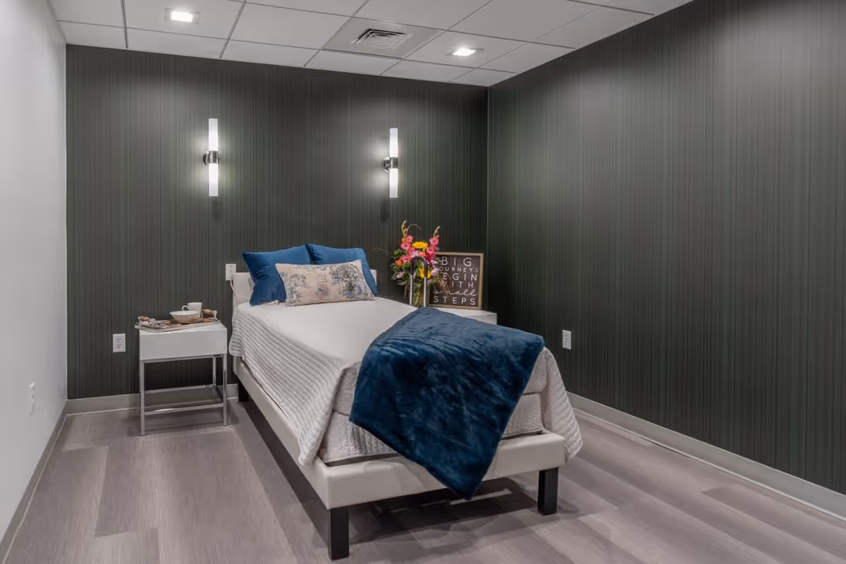A small, neatly arranged bedroom with a single bed covered in white bedding and blue pillows and blanket. There are two modern wall lights above the bed, a small white nightstand with a tray holding a cup and bowl, and a second nightstand with a colorful flower arrangement and a framed quote that reads 'Big journeys begin with small steps'. The walls are dark green with subtle vertical stripes, and the floor is light gray wood.