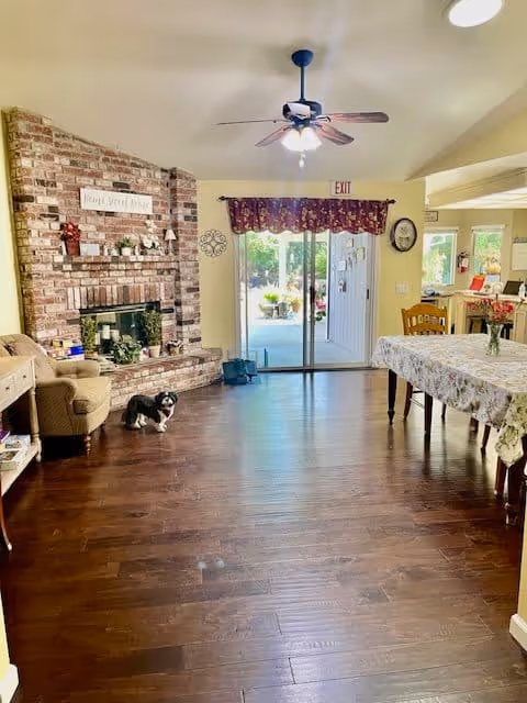A cozy senior living common area with a brick fireplace on the left, a small dog sitting on the wooden floor near an armchair, a ceiling fan with lights, and a dining table covered with a floral tablecloth on the right. Sliding glass doors with a floral valance lead to an outdoor area, and the room is warmly lit with natural light.