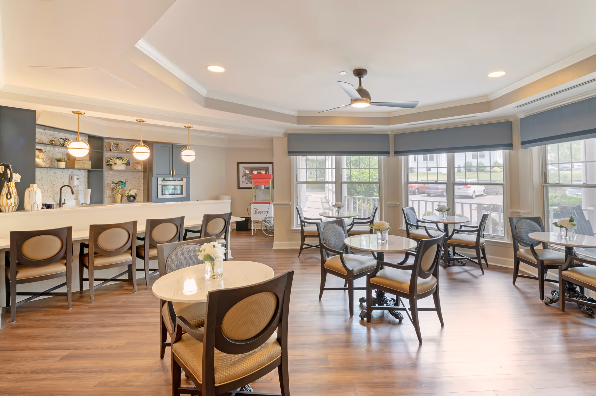 Bright communal dining room with round tables and chairs, a long serving bar with pendant lights, and large windows.