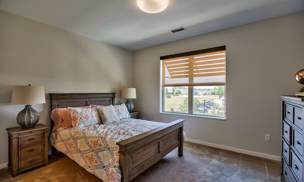 A bedroom with a wooden bed frame and a patterned bedspread with multiple pillows. There are two matching wooden nightstands on either side of the bed, each with a lamp. A window with a beige roller shade lets in natural light, and a wooden dresser is visible on the right side of the room. The walls are painted light beige and the carpet is a neutral tone.