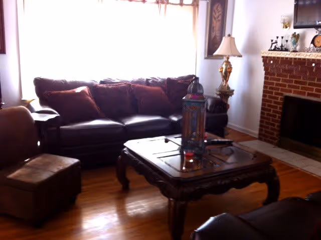 A cozy living room with a dark leather sofa adorned with several cushions, a wooden coffee table with decorative items on it, a side chair, a floor lamp with a decorative base, and a brick fireplace with a mantle holding various ornaments. The room has wooden flooring and a large window with sheer curtains letting in natural light.