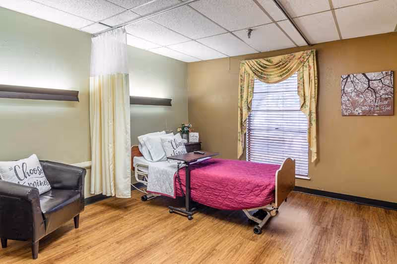 A senior living facility room with a hospital-style bed covered in a magenta quilt and white pillows, one pillow has the words 'Choose Happiness'. There is a black leather armchair with a matching pillow, a small table with flowers and a sign, a window with floral curtains and blinds, and a wall decoration with a tree and text. The floor is wooden and the walls are painted in light green and beige tones.