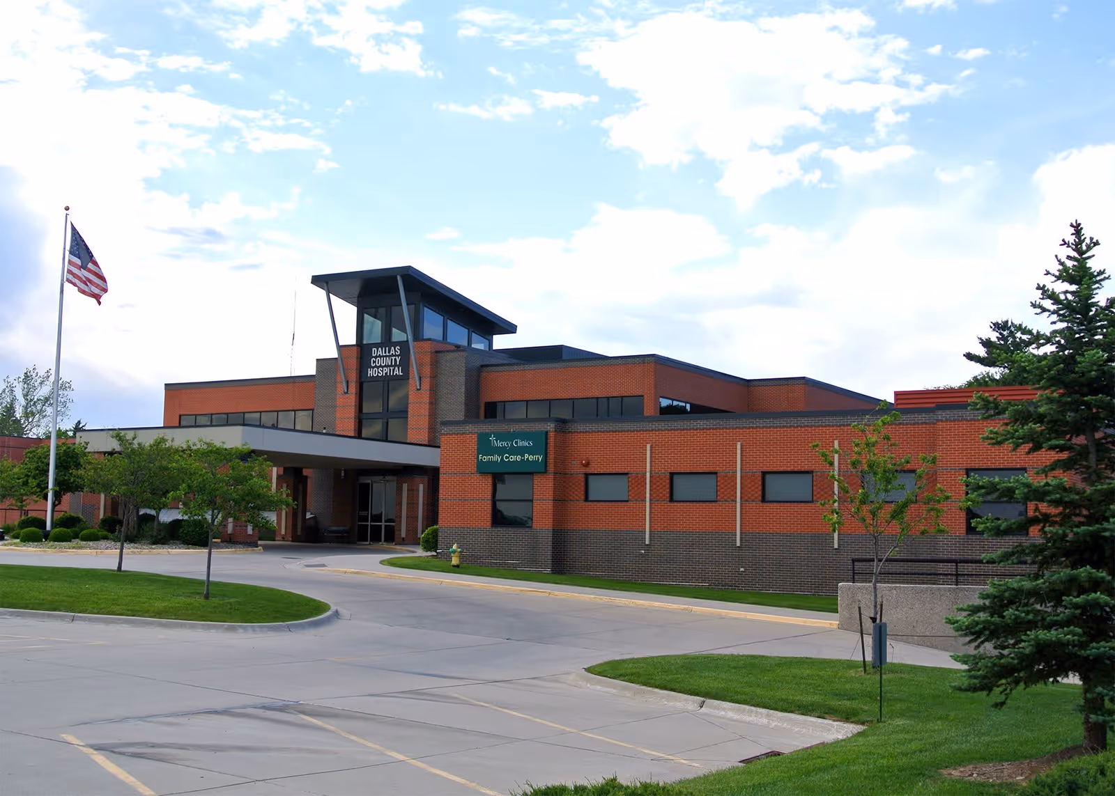 Exterior view of a modern brick building with a sign that reads Dallas County Hospital and Mercy Clinics Family Care-Perry. There is a driveway and parking area in front, a flagpole with an American flag, and some trees and grass landscaping around the building.
