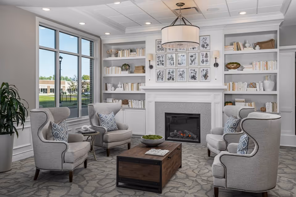 A cozy living room area with four gray upholstered wingback chairs arranged around a wooden coffee table. The room features a fireplace with a white mantel, above which are framed botanical prints. Built-in white bookshelves flank the fireplace, filled with books and decorative items. A large window lets in natural light, and a modern drum pendant light hangs from the ceiling.