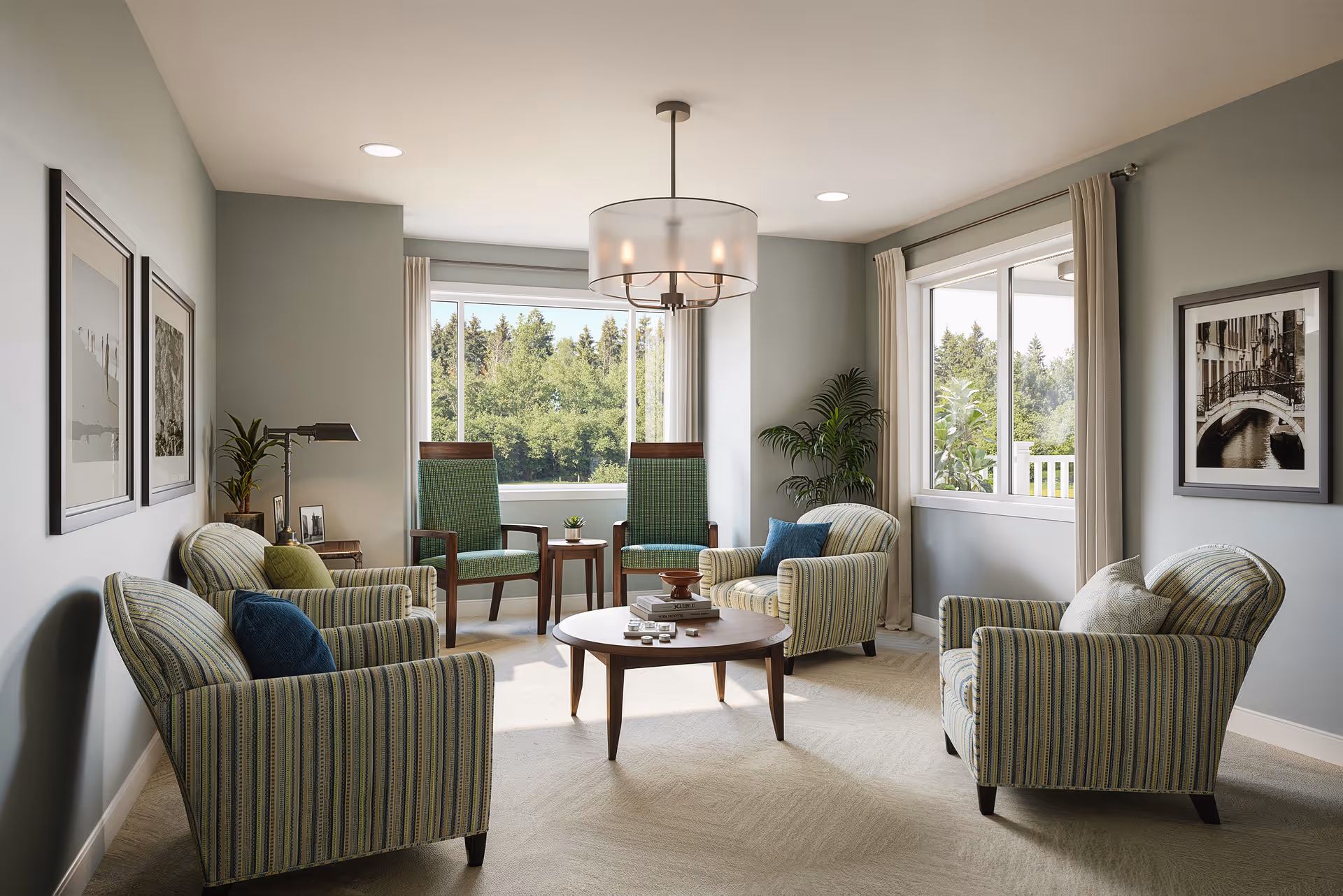 A bright and cozy living room with four striped armchairs arranged around a round wooden coffee table. Two green wooden chairs with armrests are placed near a large window showing green trees outside. The walls are painted light gray and decorated with framed black and white photographs. A modern ceiling light fixture hangs above the seating area, and there are plants and a small side table with a lamp in the corner.