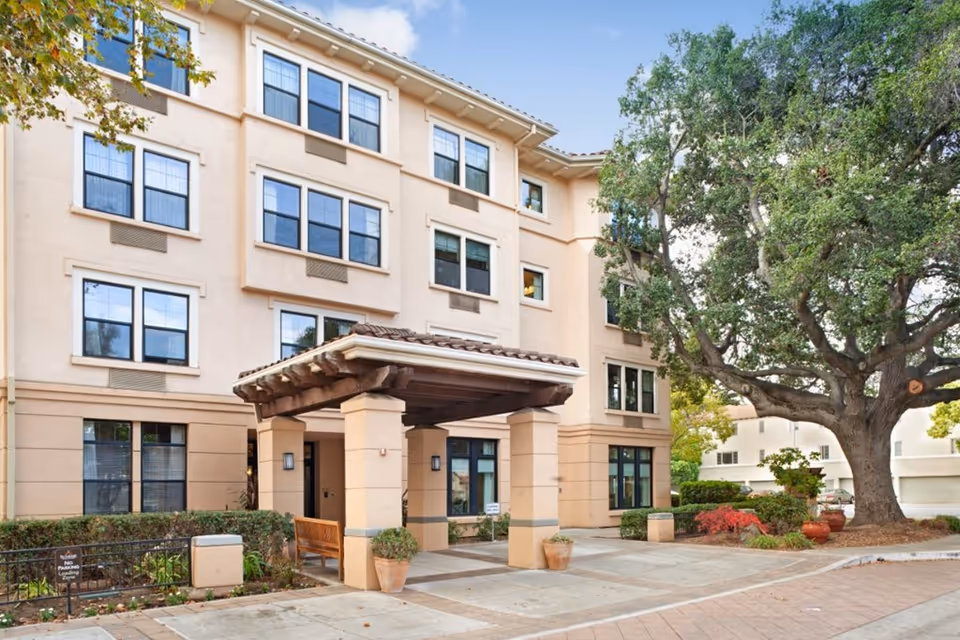 Exterior view of a four-story beige senior living facility building with multiple windows, a covered entrance supported by columns, a bench, potted plants, and a large tree on the right side.