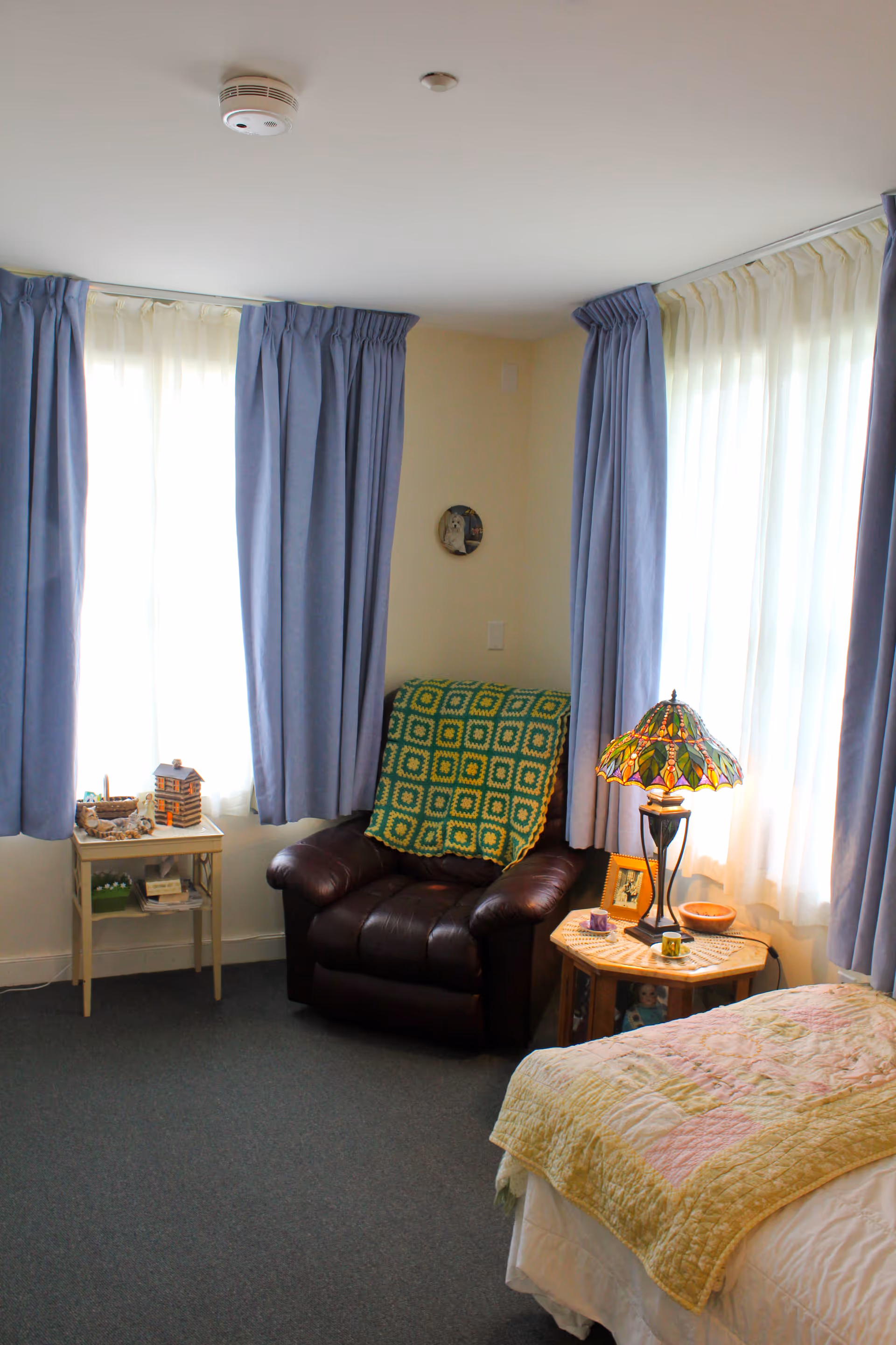 A cozy bedroom corner with a dark brown leather armchair draped with a green and yellow crocheted blanket, next to a wooden side table with a colorful stained glass lamp, a framed photo, and small decorative items. The room has light blue curtains over two windows and a bed with a yellow and pink quilt partially visible.