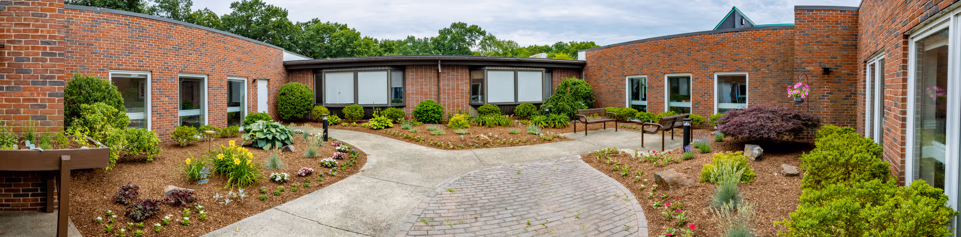 A landscaped outdoor courtyard area surrounded by a single-story brick building with multiple windows. The courtyard features paved walkways, garden beds with various plants and shrubs, benches, and a green lawn area under a partly cloudy sky.