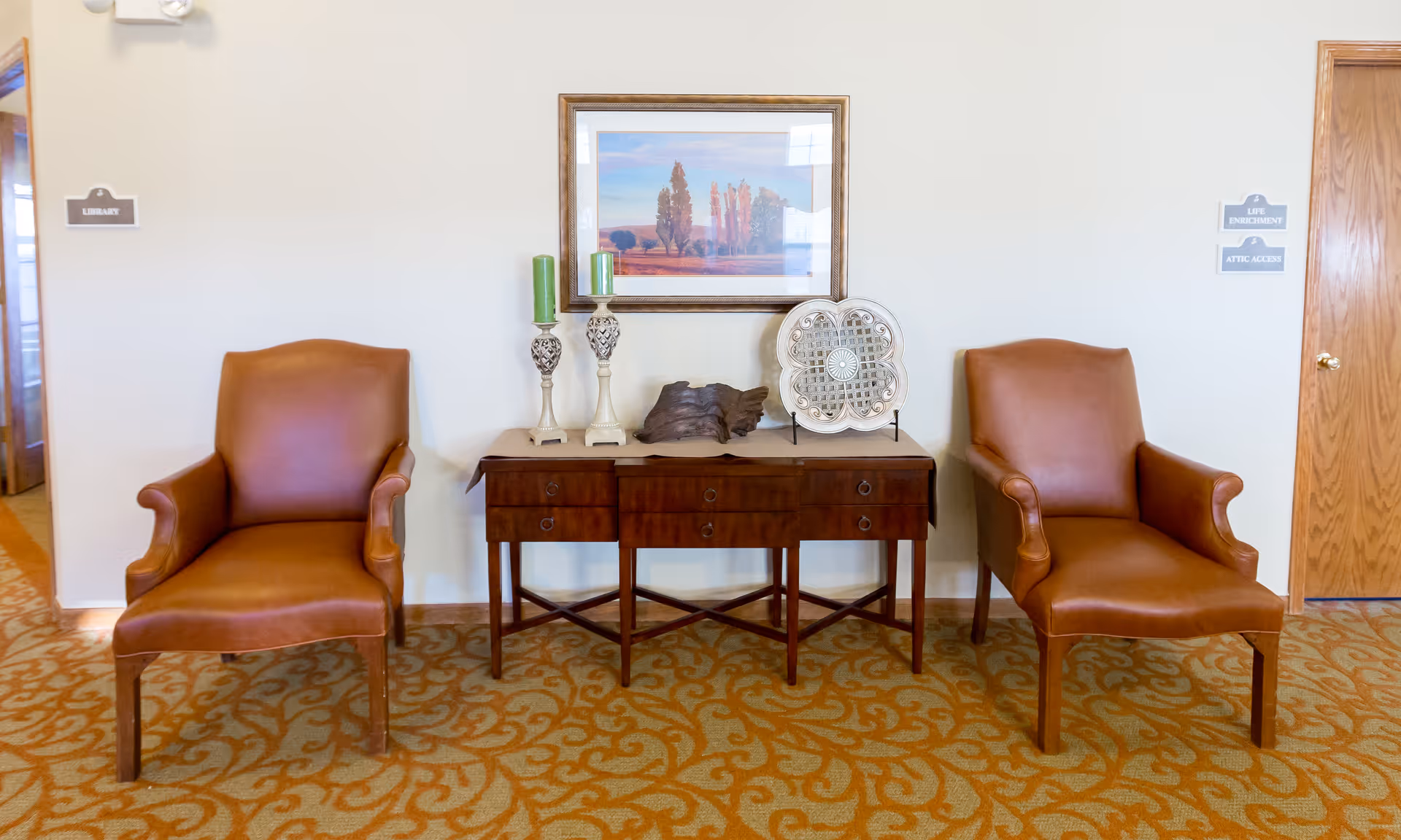 Two brown leather armchairs flanking a wooden console table topped with decorative items and a framed landscape on a carpeted lounge wall.