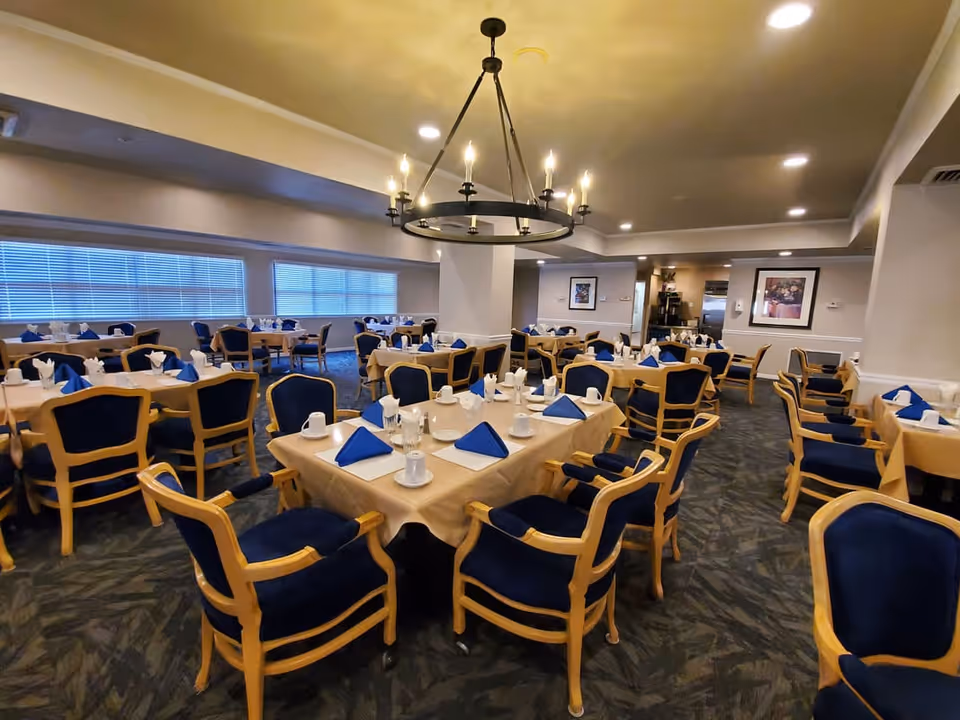 A spacious dining room with multiple tables covered in beige tablecloths, each set with white cups, plates, and blue folded napkins. The chairs have wooden frames with blue cushions. The room is well-lit with ceiling lights and a large circular chandelier in the center. Windows with blinds line one wall, and framed artwork is visible on the walls.
