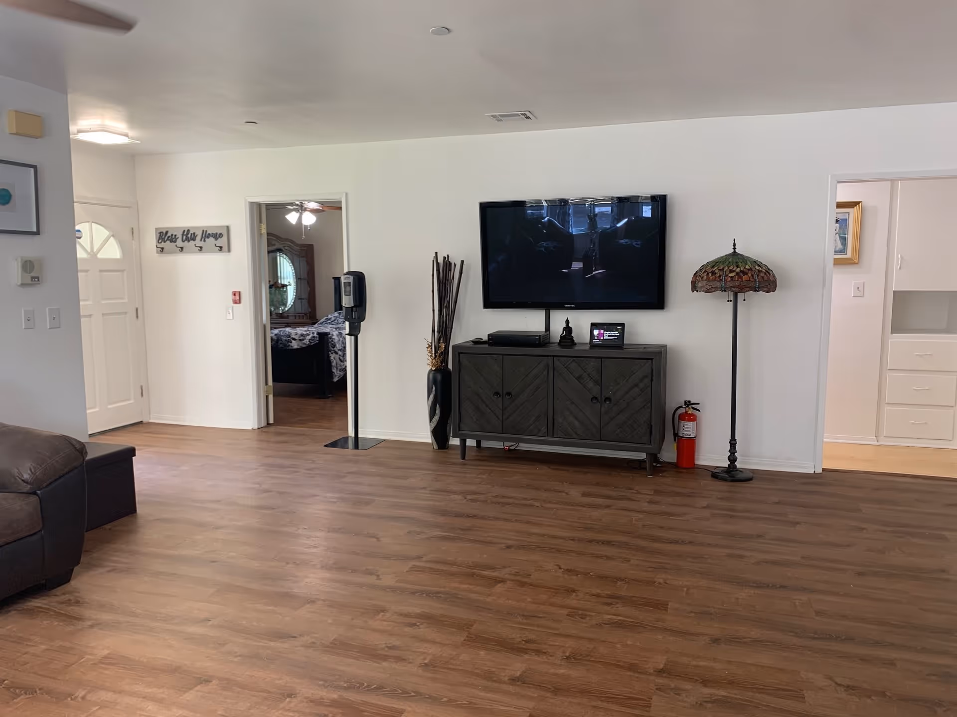 A spacious living room with wooden flooring, a dark brown leather couch on the left, a wall-mounted flat-screen TV above a dark wooden cabinet, a decorative floor lamp with a stained glass shade, a fire extinguisher, and a hand sanitizer dispenser. Two doorways lead to other rooms, one showing a bedroom with a bed and dresser. A sign on the wall reads 'Bless this Home.'