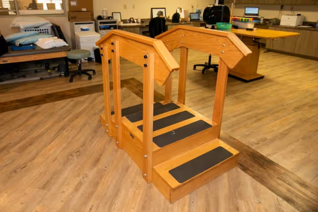 A wooden rehabilitation step platform with handrails on both sides, placed on a wood-patterned floor in a therapy or rehabilitation room. In the background, there are office desks, chairs, shelves, and various equipment and supplies.