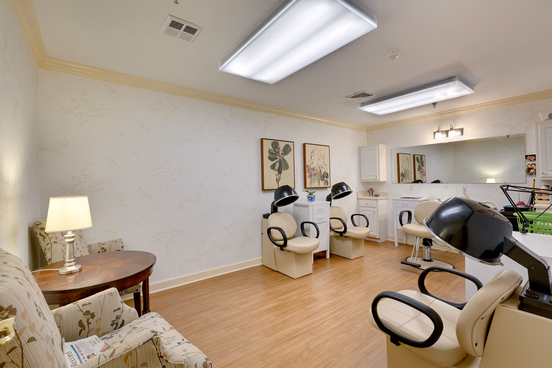 Interior view of a salon room in a senior living facility with beige salon chairs equipped with hair dryers, a wooden side table with a lamp, floral patterned armchairs, framed botanical artwork on the wall, and a large mirror above a white counter with cabinets.