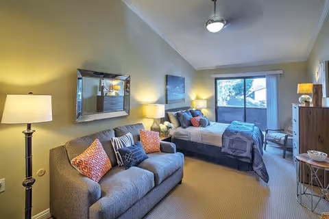 A cozy senior living bedroom featuring a gray sofa with colorful pillows, a bed with gray and blue bedding, bedside tables with lamps, a large mirror on the wall, a ceiling fan, and a sliding glass door leading to an outdoor balcony.