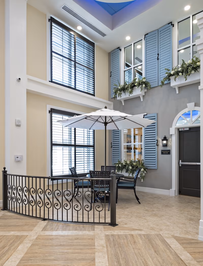 Indoor seating area with a round table and four chairs under a large white umbrella, surrounded by a decorative black metal railing. The space features tall windows with black shutters, light beige walls, and a gray wall with blue window shutters and flower boxes. A door labeled 'Magnolia House' is visible on the right side.