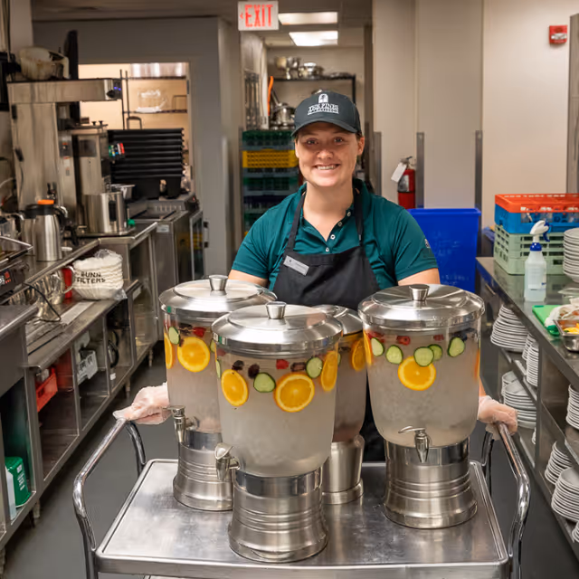 A smiling staff member in a commercial kitchen pushes a cart holding three fruit-infused water dispensers.