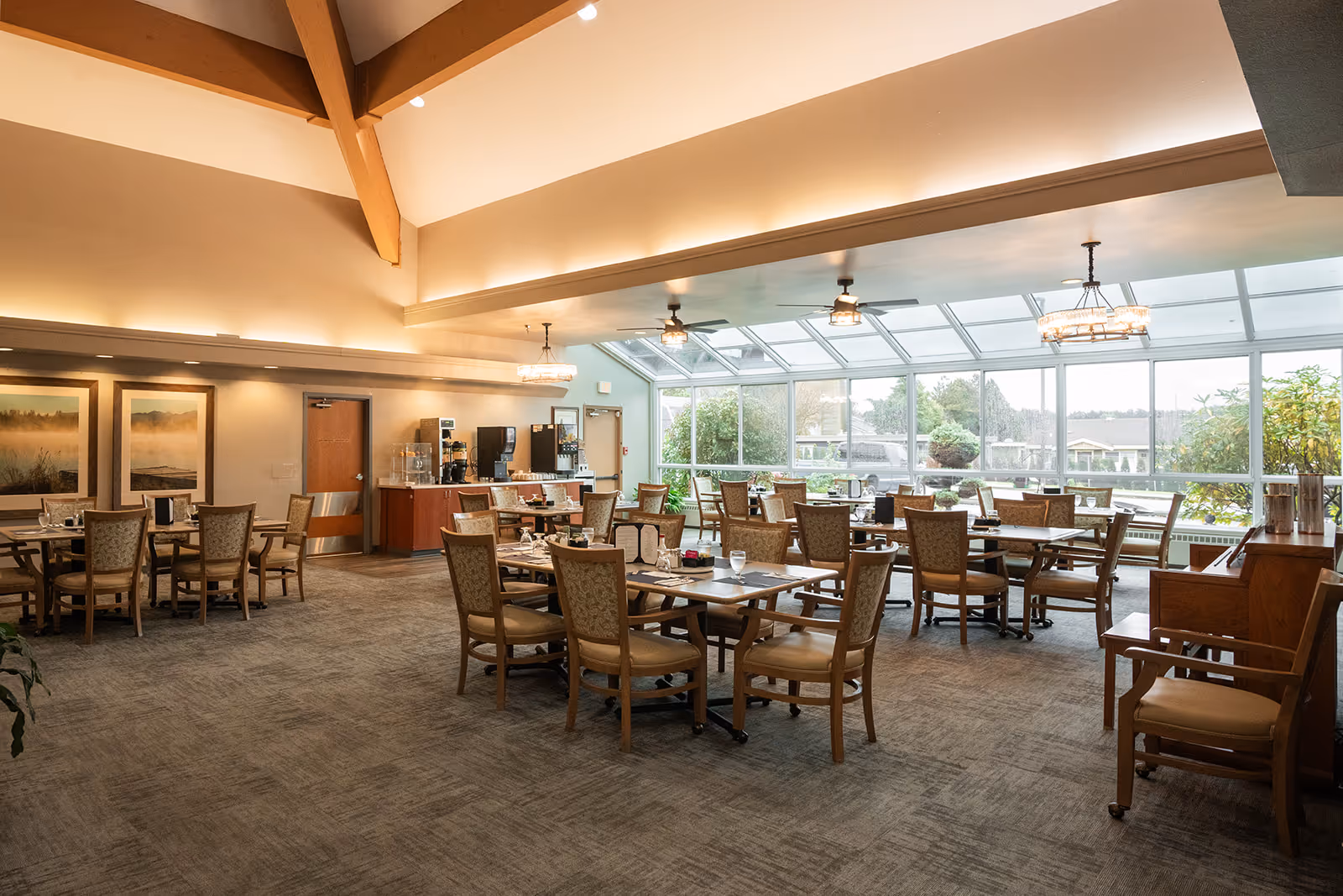 A spacious dining room with multiple wooden tables and chairs arranged neatly. The room features large windows along one side, allowing natural light to fill the space and offering a view of greenery outside. The ceiling has exposed wooden beams and modern light fixtures. There is a beverage station with coffee machines and water dispensers against one wall.