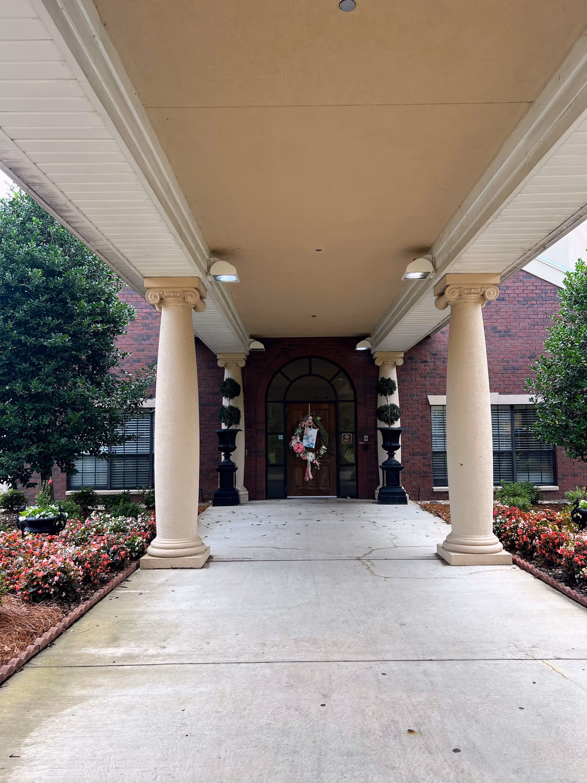 Covered portico with classical columns leading to a brick building entrance with double doors decorated by a wreath and flanked by planters and flowers.
