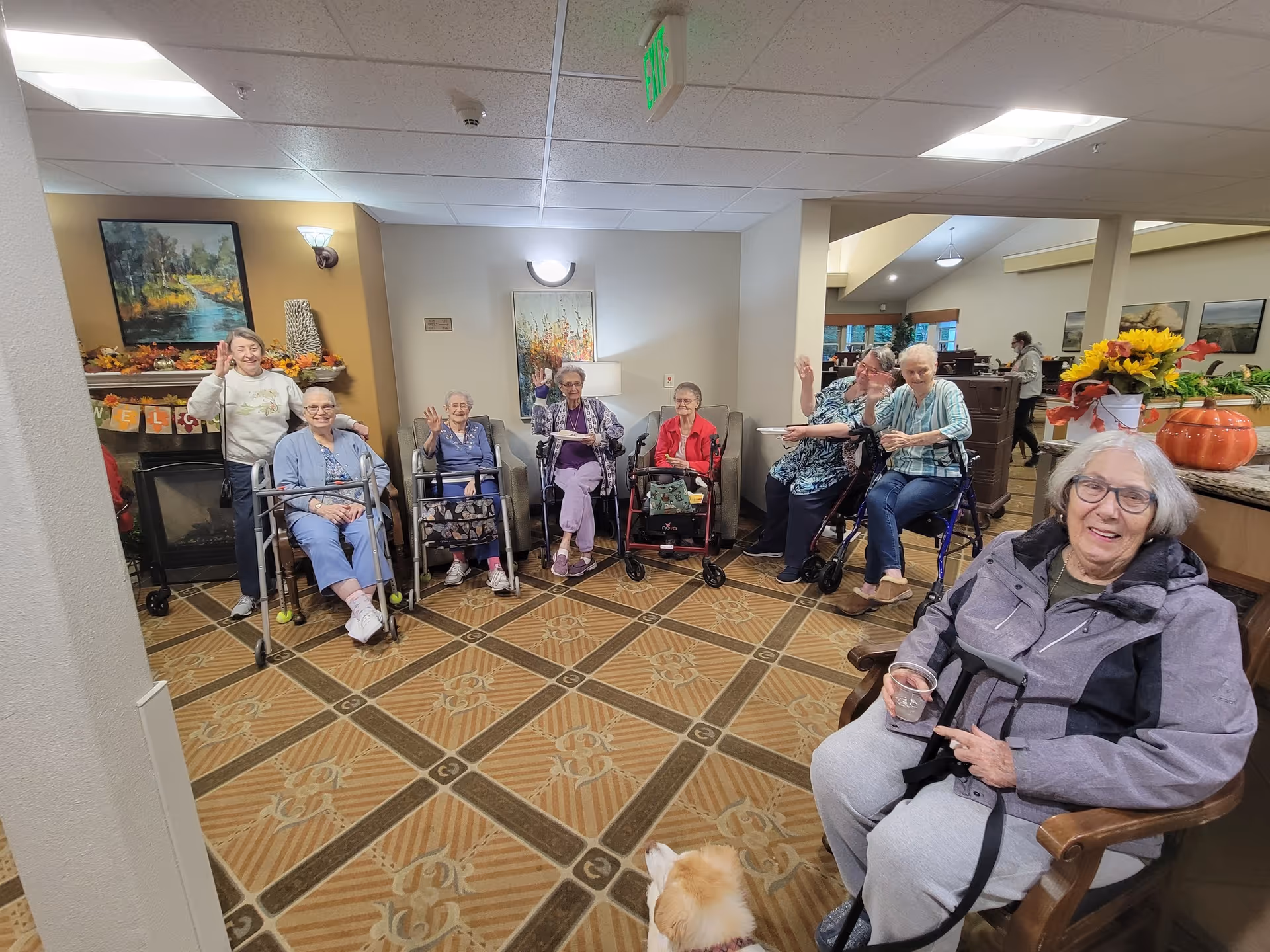 A group of elderly women sitting and standing in a common area of a senior living facility. Some are using walkers and wheelchairs. They are smiling and waving at the camera. The room has warm lighting, a patterned carpet, and autumn-themed decorations including a pumpkin and a 'Welcome' banner on a fireplace mantel.