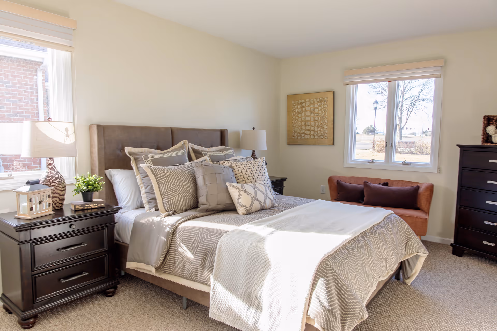 A bright and cozy bedroom with a large bed adorned with multiple decorative pillows and a beige patterned bedspread. On either side of the bed are dark wooden nightstands with lamps, a small plant, and decorative items. There are two windows letting in natural light, a framed artwork on the wall, a small brown loveseat under one window, and a dark wooden dresser on the right side of the room.