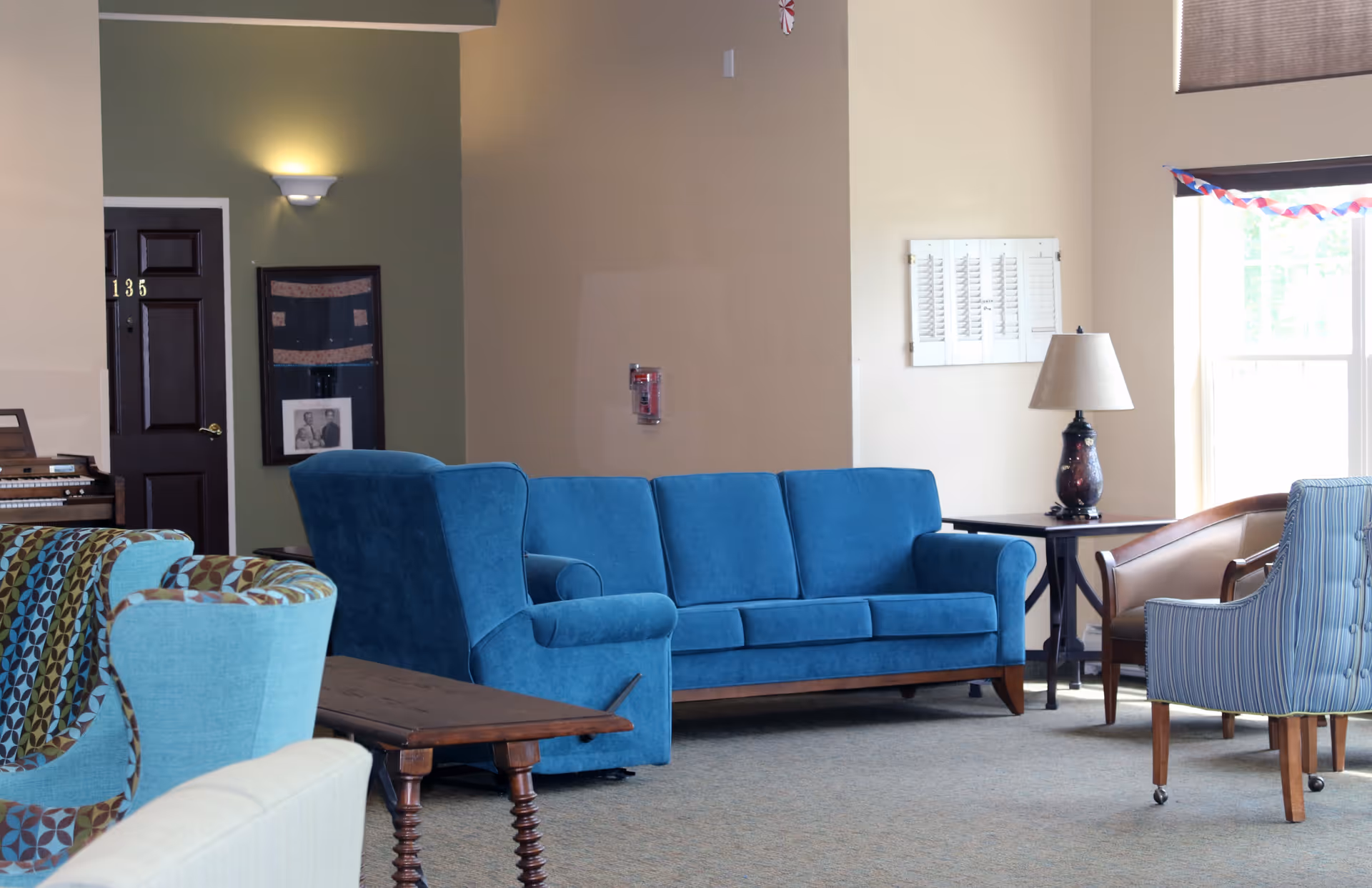 A cozy living room area with a blue sofa, a blue armchair, a patterned armchair, a wooden side table, and a lamp on a small table near a window with blinds. The walls are painted beige and green, and there is a door numbered 135 in the background along with a framed picture and a fire alarm on the wall.