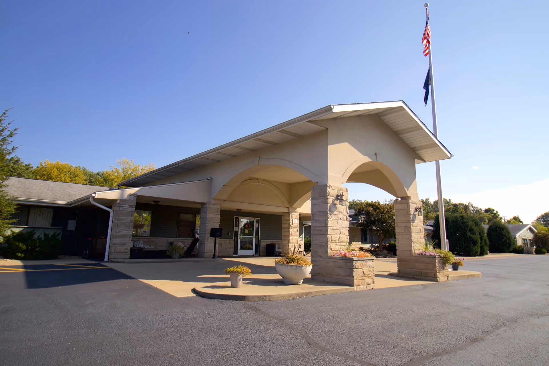 Exterior view of the entrance to Majestic Care of Bedford, featuring a covered driveway with stone pillars, potted plants, and an American flag on a flagpole against a clear blue sky.