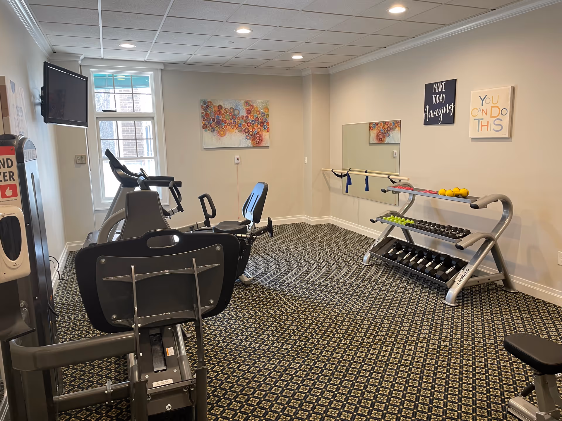 A small fitness room with exercise equipment including a recumbent bike and treadmill, a rack of various dumbbells, a wall mirror, and motivational wall art that says 'Make Today Amazing' and 'You Can Do This'. There is a window letting in natural light and a hand sanitizer dispenser mounted on the left wall.