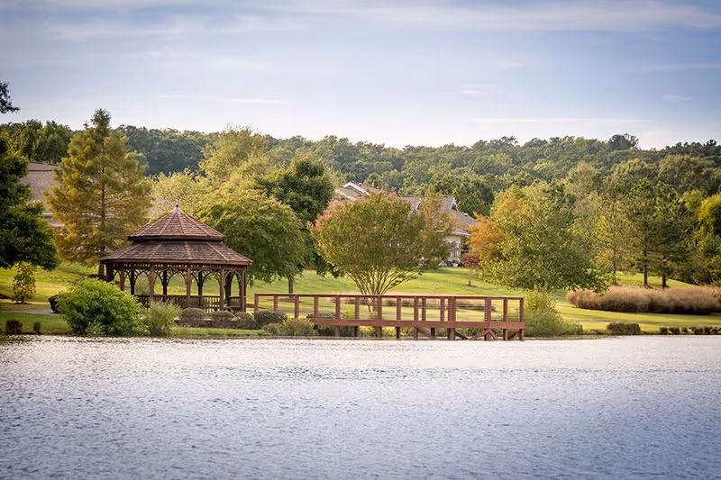 A peaceful outdoor scene at Croasdaile Village featuring a wooden gazebo and a wooden bridge over a body of water, surrounded by green trees and grassy areas under a partly cloudy sky.