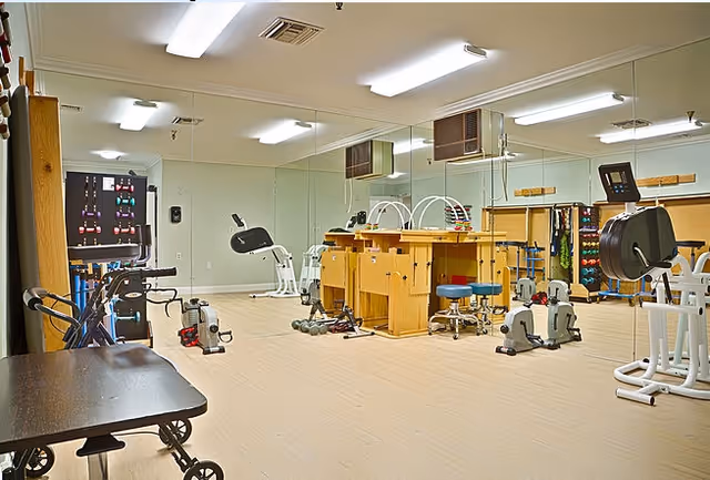 A well-lit physical therapy or rehabilitation room with various exercise equipment including stationary bikes, weights, and therapy tables. The room has large mirrors on one wall and light-colored flooring.