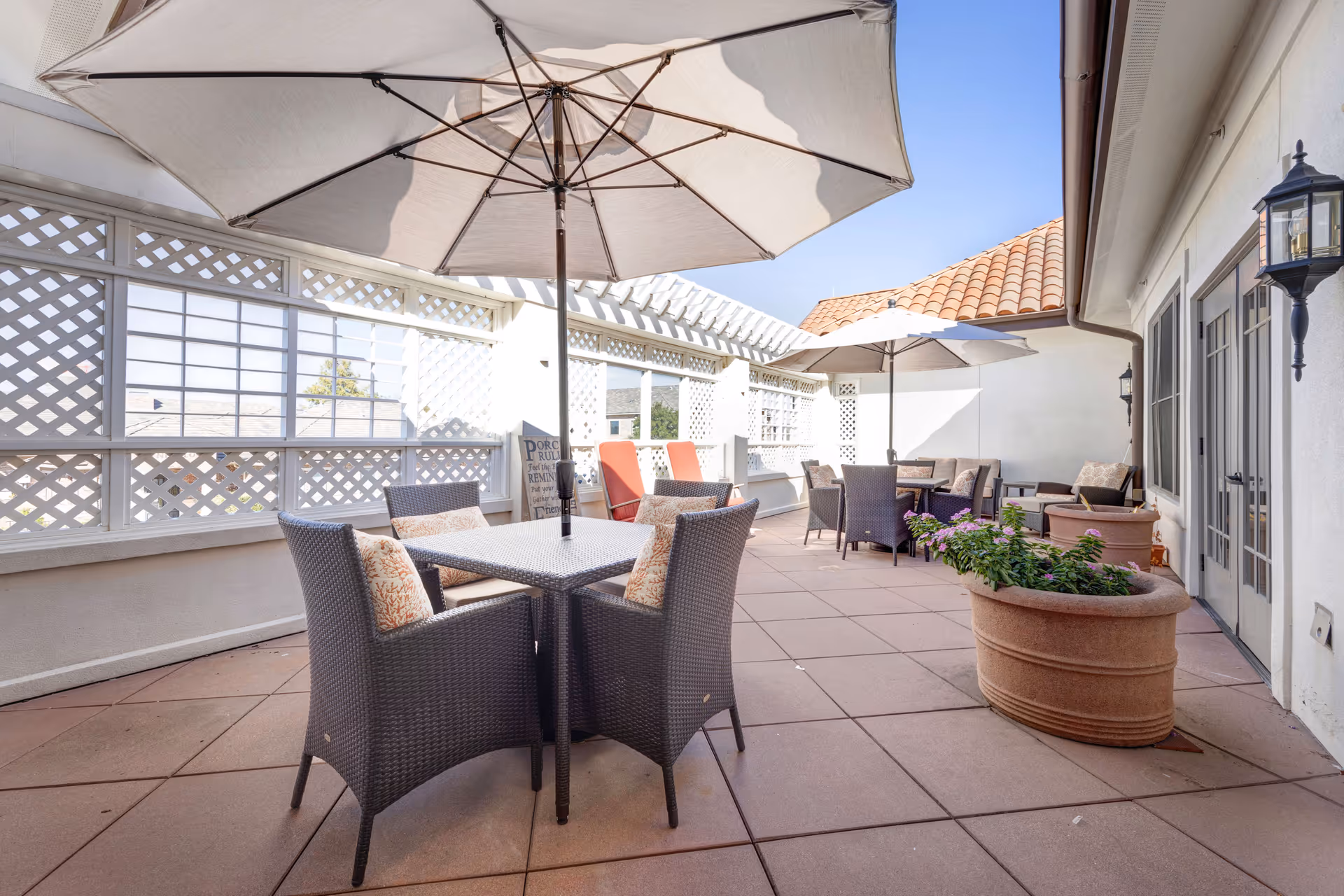 Outdoor patio area with several tables and chairs under large umbrellas, surrounded by white lattice fencing and potted plants, with a tiled roof visible in the background.