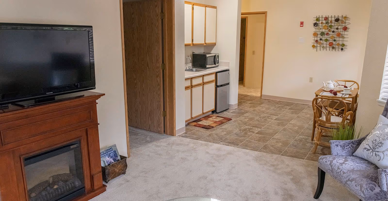 Interior view of a senior living facility unit showing a living area with a TV on a wooden stand with a fireplace, a small kitchenette with a microwave and mini fridge, a dining table with two chairs set for tea, and a decorative wall piece. The floor transitions from carpet in the living area to tile in the kitchenette and dining area.