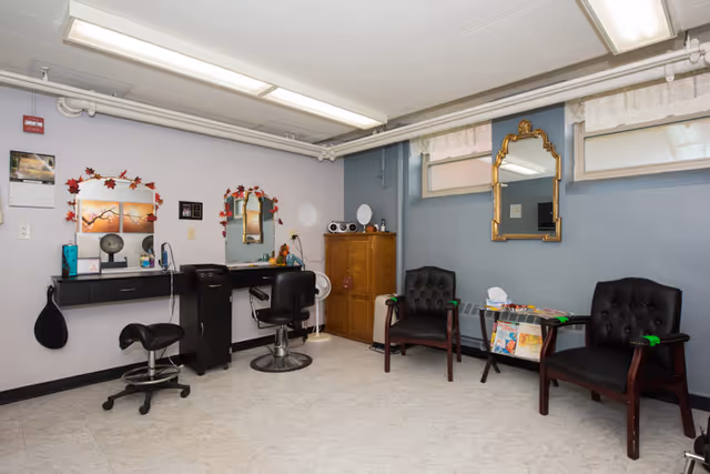 A small salon area in a rehabilitation and nursing center with two black salon chairs in front of mirrors on the left side, a wooden cabinet in the corner, and two black cushioned chairs with armrests on the right side next to a small table holding magazines. The walls are painted light blue with two small windows near the ceiling and a decorative gold-framed mirror between them.