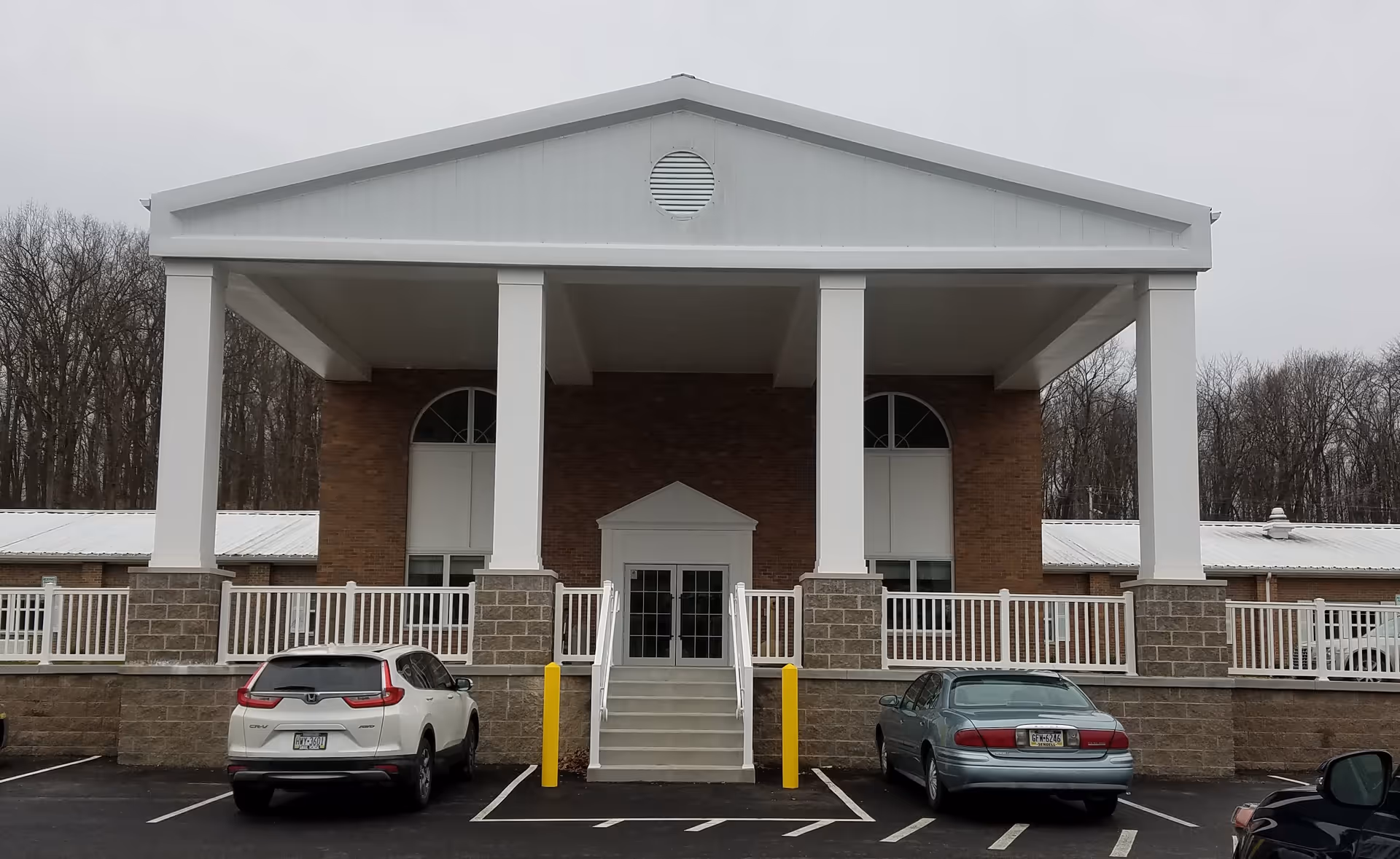 Front exterior view of a brick building with a large white portico supported by four white columns. There are two cars parked in front of the building, and a set of stairs leads up to double glass doors. The sky is overcast and there are leafless trees in the background.
